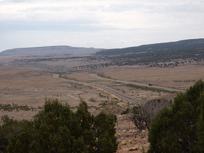 [The two separate roadways (eastbound and westbound) extending into the distance over mostly flat land. There are some trees in the foreground (at the top of the hill from which this photo was taken) and one far distant sloping hillside is also covered with dark green shrubbery.]